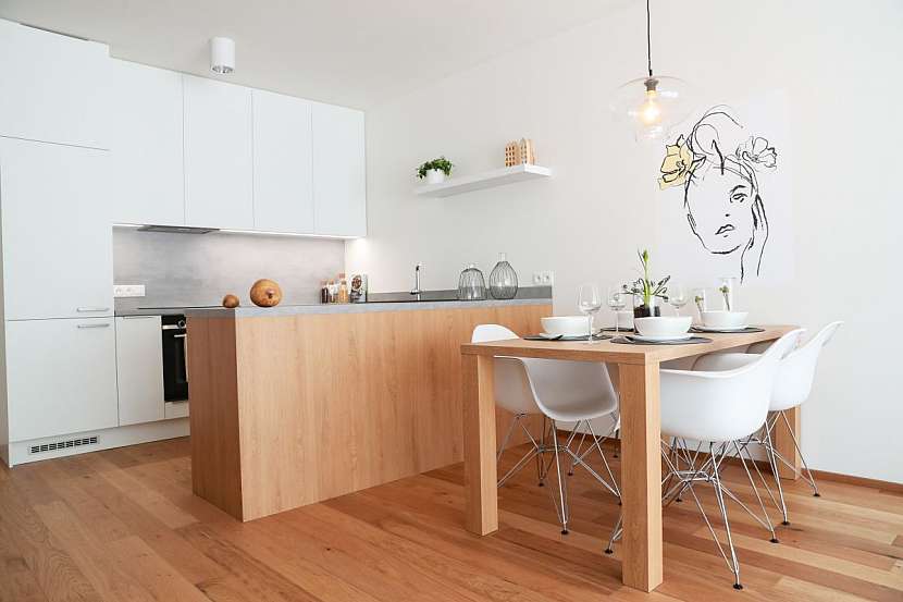 Obývací pokoj vzorového bytu je propojen s kuchyňským koutem, v němž je umístěna kuchyňská linka ve tvaru písmene L v provedení bílá a dub.