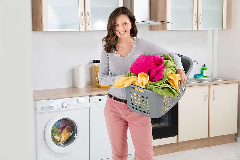 Žena v kuchyni u pračky s plným košem barevného prádla