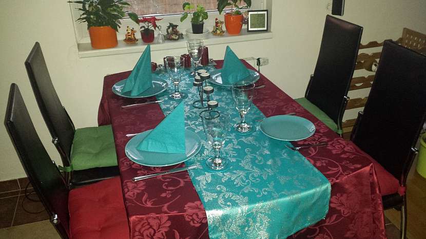 Slavnostně vyzdobený jídelní stůl je opatřen vzorovaným barevným látkovým ubrusem s hedvábným leskem a na něm je umístěn barevný středový vánoční látkový ubrus rovněž se vzorem, který má třpytivý stříbrný nádech. Jídelní set i papírové ubrousky jsou zvoleny v barvě středového ubrusu. Rovněž je ve stejné barvě s velkým ubrusem adventní věnec, který je v moderním stylu
