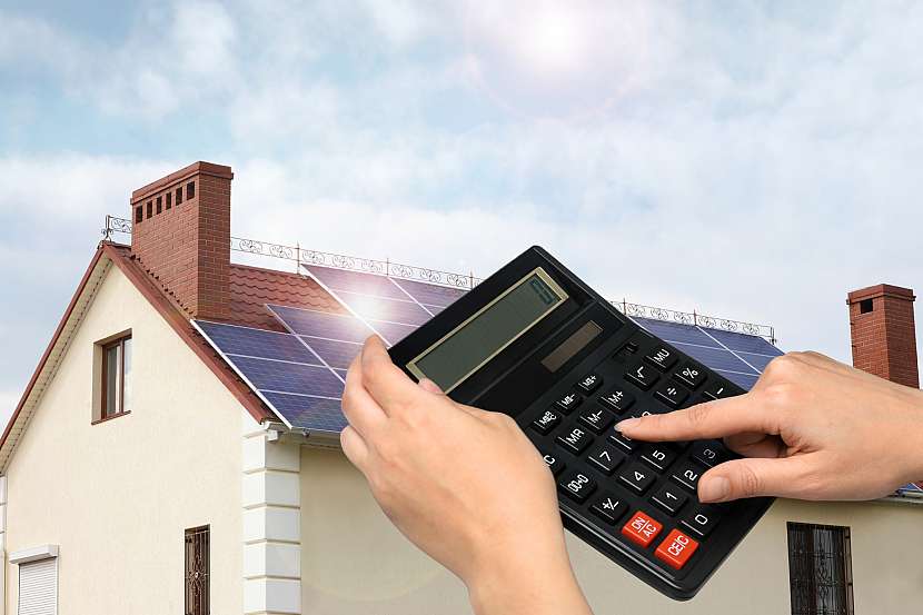 Ruka s kalkulačkou, v pozadí dům s fotovoltaikou